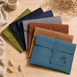 May include: A collection of rectangular document folders in various colors including olive green, black, navy blue, gray, tan, brown, and blue. Each folder features a flap with a vertical strap closure. The blue folder has a ginkgo leaf graphic and the word Serene.