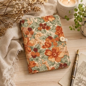 May include: A rectangular notebook with a textured fabric cover featuring a floral pattern in shades of orange, rust, and sage green. The cover is secured with a small, round, cream-colored button and a loop closure on the right side.