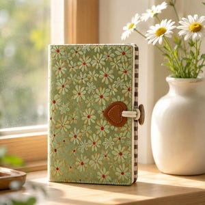Può includere: Un diario verde salvia con un motivo ricamato di margherite bianche con centri rossi. La copertina include una chiusura con bottone marrone a forma di cuore con un passante elastico beige e un dettaglio del dorso in tessuto a quadri marrone e bianco.