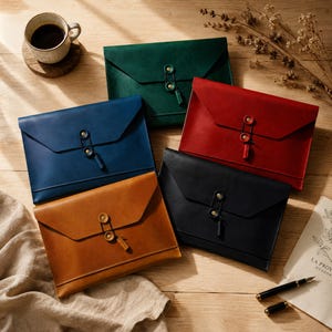 May include: Five rectangular leather document folders in green, blue, red, black, and tan. Each folder features a pointed flap secured by a brass-coloured button and elastic cord closure. The folders are arranged on a wooden surface with a coffee mug.