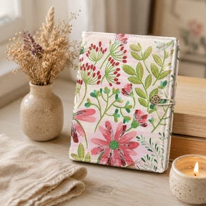 May include: A fabric-covered notebook featuring a light pink background with a vibrant botanical print of red flowers, green leaves, and delicate stems. The cover is secured with a small, circular, decorative button and a thin elastic loop closure.