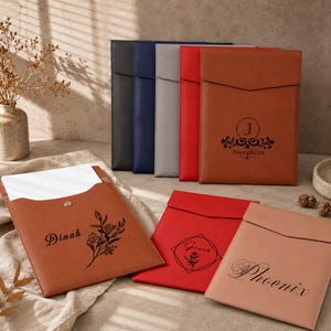 May include: A collection of rectangular document folders in black, navy, gray, red, and tan leather-like material. Each folder features a flap closure and personalized black text or floral designs including the names Dinah, Josephine, Grace, and Phoenix.