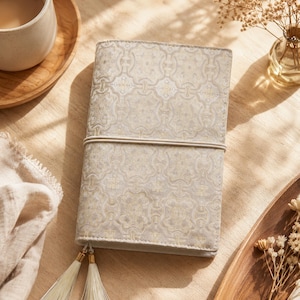 May include: A rectangular notebook cover featuring a light beige and gold intricate geometric pattern. The cover is secured with a thin elastic band wrapped horizontally around the center and includes two dangling off-white tassels at the bottom.
