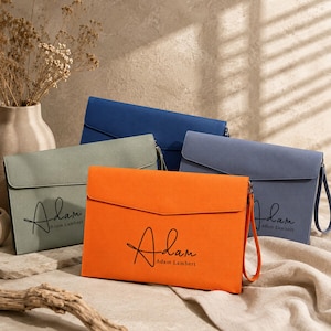 May include: A collection of four rectangular envelope-style clutch bags in orange, sage green, dark blue, and light blue. Each bag features a flap closure, a wrist strap, and the black printed text Adam Lambert in a cursive and sans-serif font.