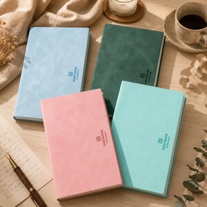 Puede incluir: Cuatro cuadernos rectangulares con cubiertas mate de colores sólidos en azul claro, verde oscuro, rosa y verde menta. Cada cuaderno presenta un pequeño icono de cuadrícula y el texto "GRID BOOK A5 Note Series" impreso en color oscuro.