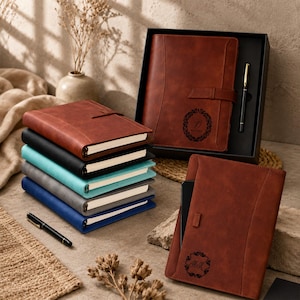 Op de afbeelding: Een verzameling rechthoekige notitieboeken met gladde covers in bruin, zwart, taling, grijs en blauw. Eén bruin notitieboek zit in een zwarte geschenkdoos met een pen. Een ander bruin notitieboek heeft een cirkelvormig monogram met de letters HK.
