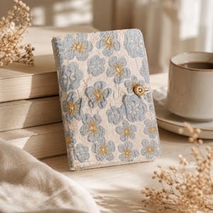 May include: A rectangular notebook cover featuring a cream-colored fabric base with an embroidered pattern of light blue flowers. Each flower has a small gold-colored center. The cover is secured with a small, round, light brown wooden button closure.