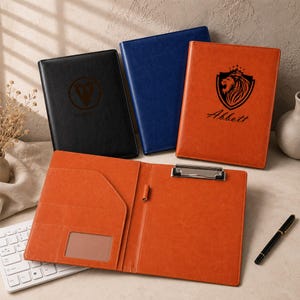May include: A collection of three rectangular portfolios in black, blue, and orange. The orange portfolio is open, revealing a metal clipboard clip, a pen loop, and a clear ID window. The orange and black portfolios feature custom logos.