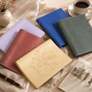 May include: A collection of five rectangular leather journals in lavender, dusty rose, yellow, blue, and dark green. The yellow journal features a delicate, engraved floral vine pattern on the front cover. Each journal has a smooth, matte finish.