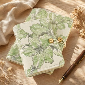 May include: Two rectangular notebooks featuring a cream-colored fabric cover with a repeating light green and metallic silver floral pattern. Each notebook is secured with a thin cord wrapped around a small, circular, gold-toned metal button.