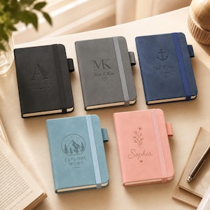 Puede incluir: Cinco cuadernos rectangulares con cierres elásticos y portabolígrafos. El cuaderno negro muestra la letra A y ALEXANDER. El gris muestra MK, Mia & Kai y un corazón. El azul muestra un ancla, Jack Wilson y 2025. El azul claro muestra un gráfico de montaña y EXPLORE MORE. El rosa muestra un diseño floral y Sophia.