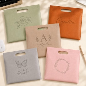 Pode incluir: Cinco sacos quadrados de feltro com pegas recortadas em verde sálvia, laranja, bege, cinzento e rosa. O saco verde apresenta uma flor, o laranja diz Camellia, o bege mostra a letra A e Amelia, o cinzento mostra uma borboleta e Lily, e o rosa tem uma coroa.