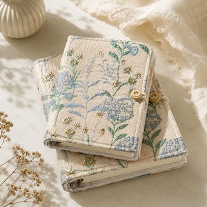May include: Two stacked notebooks with a cream-coloured fabric cover featuring an embroidered botanical pattern in shades of blue, green, and gold. Each notebook is secured with a small, round, light-coloured wooden button and a thin elastic cord.