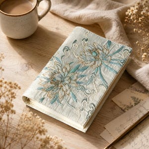 Puede incluir: Un cuaderno rectangular con una cubierta de tela azul claro que presenta un intrincado bordado de dos grandes flores en tonos azul cerceta, blanco y dorado, con patrones de enredaderas que se extienden por la superficie.