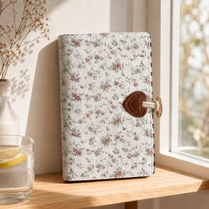 May include: A rectangular notebook with a white cover featuring a small-scale floral pattern in pink and green. The notebook is secured with a brown heart-shaped leather-like tab and a light-colored cord closure attached to the side edge.