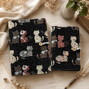 Puede incluir: Dos cuadernos cubiertos de tela con fondo negro y un patrón repetitivo de gatos en varios estampados florales y de lunares. Cada cuaderno está asegurado con un pequeño botón de madera y un cierre de cordón elástico blanco.