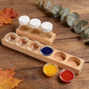 Könnte beinhalten: Holz-Malpalette mit drei kleinen Gläsern mit weißen Deckeln und fünf kleinen Glasschalen. Die Schalen enthalten blaue, gelbe und rote Farbe. Die Palette befindet sich auf einer Holzoberfläche.