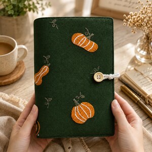 May include: A dark green fabric notebook cover featuring embroidered orange pumpkins with white detailing. The cover includes a light-coloured wooden button closure attached with a white cord, securing the front flap of the rectangular journal.