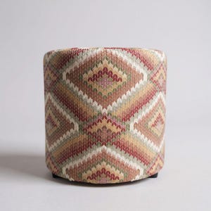 Puede incluir: Un otomán cilíndrico con un patrón geométrico en tonos rojos, beige, verdes y blancos. El taburete cubierto de tela tiene una base resistente y es adecuado para sentarse o como elemento decorativo.
