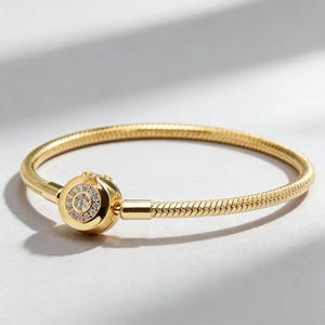 May include: A gold bracelet with a snake chain design. The bracelet has a round clasp with a circle of small, clear gemstones. The bracelet is displayed on a white surface.
