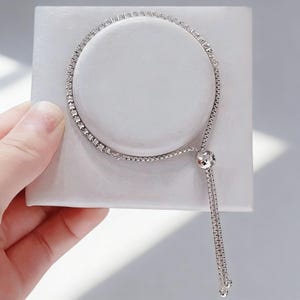 May include: A silver bracelet with a row of small, clear stones set along the top. The bracelet has a delicate chain and a dangling strand with small, round accents. The bracelet is displayed on a white surface.