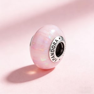 May include: A light pink, opal-like bead with a silver-tone metal band. The bead has a pearlescent effect and the word "PANDORA" is engraved on the metal band. The bead is a jewelry item, likely a charm or pendant.