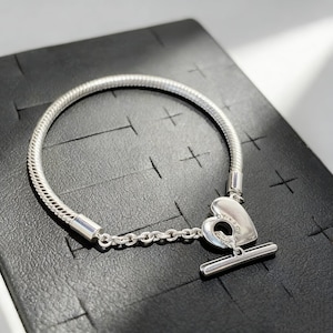 May include: A silver bracelet with a heart-shaped clasp. The bracelet is made of a snake chain design and features a T-bar closure. The heart clasp has a small loop and is connected to the bracelet with a small chain.