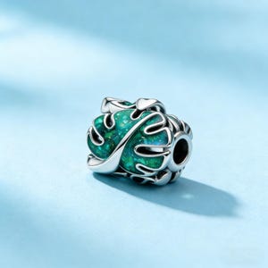 May include: A silver charm with a green, leaf-shaped design. The charm has a detailed, openwork pattern and a hole for threading onto a bracelet or necklace. The green colour has a shimmering effect.