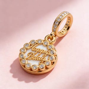 May include: A gold-coloured charm with the words "Happy Bday" in cursive on a white background. The charm is round and surrounded by small, clear gemstones. The top loop is also adorned with gemstones.