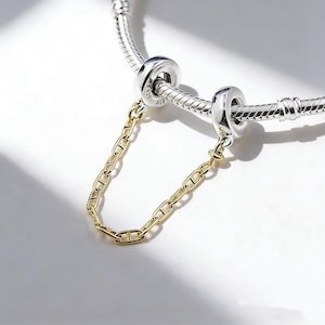 Può includere: Un bracciale d'argento con due charm bead argentati e una catena di sicurezza dorata. La catena è attaccata ai charm bead. Il bracciale è su sfondo bianco.