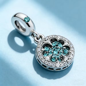 May include: A silver charm with a clover design. The charm features a cluster of teal-coloured stones in the centre, surrounded by a circle of clear stones. A teal stone is also set in the loop at the top. The charm hangs from a silver loop.