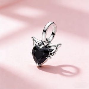 Op de afbeelding: Een zilverkleurige bedel met een zwarte hartvormige edelsteen en vleugelaccenten. De bedel heeft een lus om aan een armband of ketting te bevestigen. Het hart is facetgeslepen en reflecteert licht. De vleugels zijn gedetailleerd.