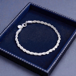 Puede incluir: Un brazalete de cadena de cuerda plateada exhibido en una caja de joyería azul oscuro. El brazalete tiene un diseño trenzado texturizado y un cierre seguro. La caja tiene un interior de terciopelo azul oscuro y un exterior azul oscuro.