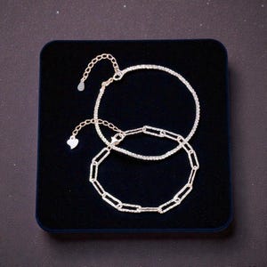 May include: Two silver bracelets displayed on a dark blue velvet surface. One is a delicate chain of small, sparkling stones. The other features a chain link design. Both have adjustable clasps.