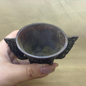 Op de afbeelding: Een kleine, antieke metalen beker met een donkere, verouderde afwerking. De beker heeft twee gestileerde vleugelvormige handvatten aan weerszijden. De binnenkant van de beker is donker en het algehele ontwerp suggereert een vintage of historische esthetiek.