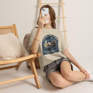 Könnte beinhalten: Ein beiges T-Shirt mit kurzen Ärmeln und einer Grafik, die eine Katze bei der Arbeit am Laptop mit einer Kaffeetasse mit der Aufschrift JAVA zeigt. Der Text lautet COFFEE CAT CODER und CODE ALL NIGHT, NAP ALL DAY, MOUSE OPTIONAL. Graue Jeansshorts.