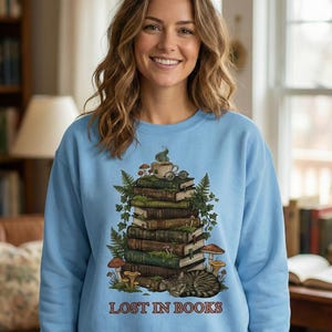 Pu&ograve; includere: Felpa azzurra con una grafica di una pila di libri, una tazza da t&egrave;, funghi e un gatto che dorme. Il testo "LOST IN BOOKS" &egrave; stampato in marrone. La felpa &egrave; a girocollo e a maniche lunghe.