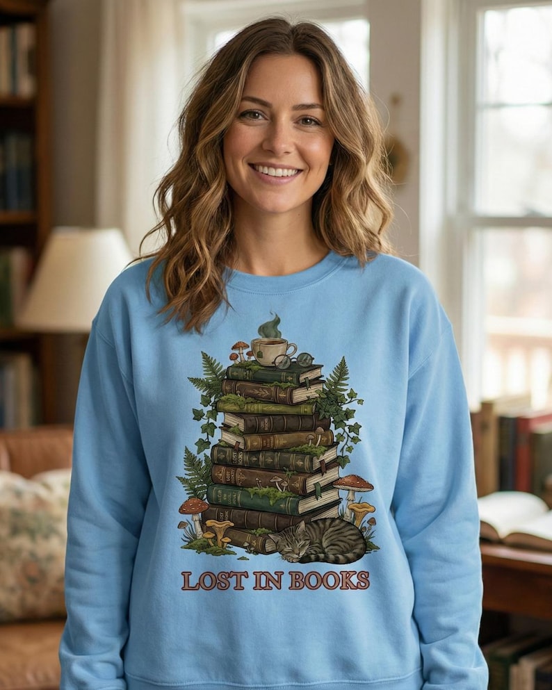 Pu&ograve; includere: Felpa azzurra con una grafica di una pila di libri, una tazza da t&egrave;, funghi e un gatto che dorme. Il testo "LOST IN BOOKS" &egrave; stampato in marrone. La felpa &egrave; a girocollo e a maniche lunghe.