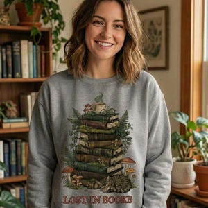 Pu&ograve; includere: Felpa grigia con un design stravagante di una pila di libri con una tazza da t&egrave;, funghi e un gatto che dorme. Le parole "LOST IN BOOKS" sono stampate in basso. La persona nell'immagine sta sorridendo.