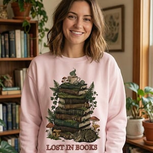 Pu&ograve; includere: Una felpa rosa chiaro con una grafica di una pila di libri, funghi e un gatto. Una tazza da t&egrave; e occhiali sono in cima ai libri. Il testo "LOST IN BOOKS" &egrave; stampato in basso. La persona nell'immagine sta sorridendo.