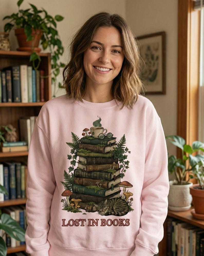 Pu&ograve; includere: Una felpa rosa chiaro con una grafica di una pila di libri, funghi e un gatto. Una tazza da t&egrave; e occhiali sono in cima ai libri. Il testo "LOST IN BOOKS" &egrave; stampato in basso. La persona nell'immagine sta sorridendo.