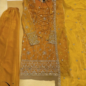 May include: Mustard yellow embroidered formal wear set. The set includes a long-sleeved tunic with intricate silver embroidery and a lace-up front, matching pants, and a sheer scarf. The outfit is embellished with sequins and beads.