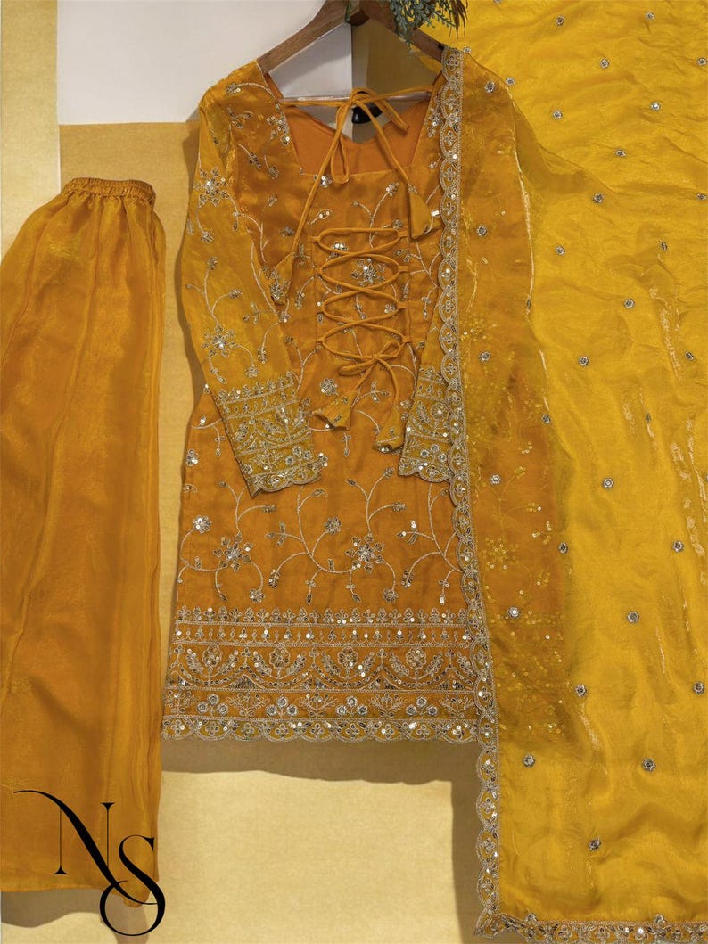 May include: Mustard yellow embroidered formal wear set. The set includes a long-sleeved tunic with intricate silver embroidery and a lace-up front, matching pants, and a sheer scarf. The outfit is embellished with sequins and beads.