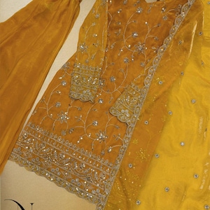 May include: Mustard yellow embroidered salwar kameez set. The ensemble includes a long-sleeved tunic with intricate silver embroidery and sequin detailing, matching pants, and a sheer dupatta. The outfit is displayed on a hanger.