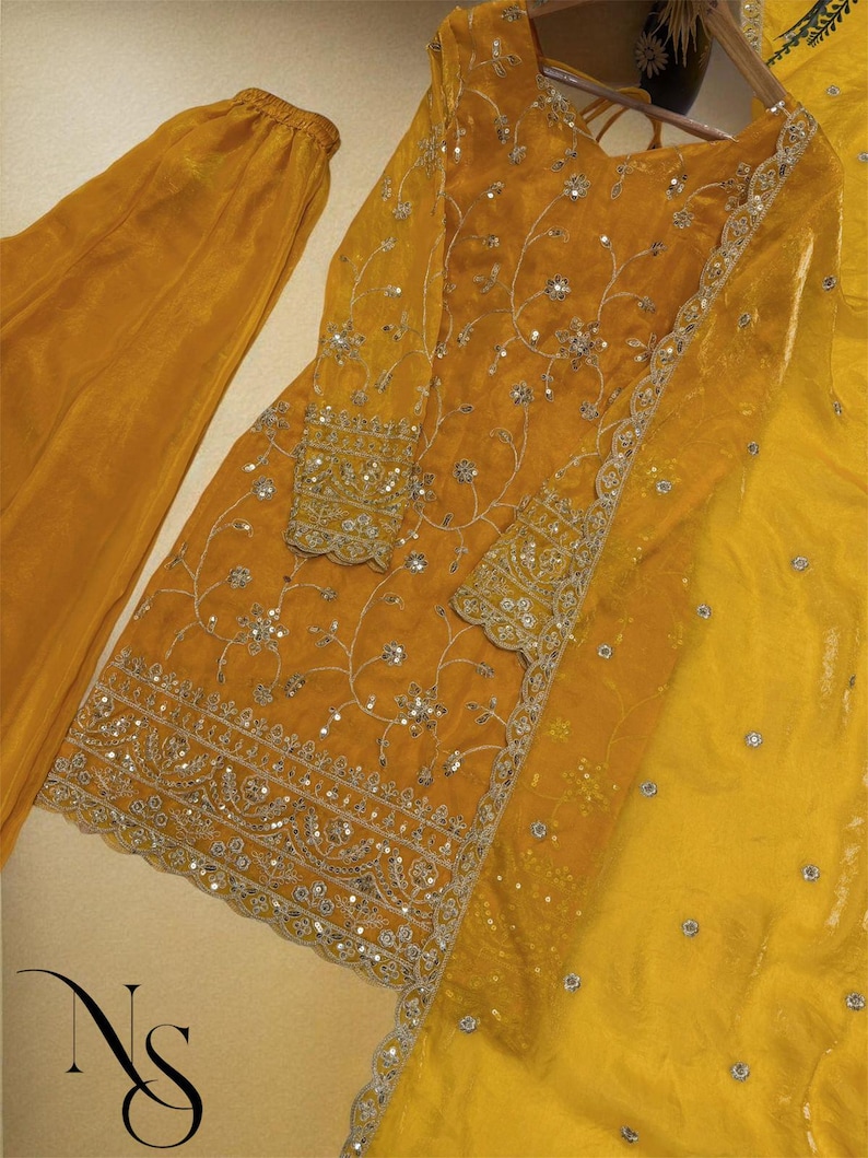 May include: Mustard yellow embroidered salwar kameez set. The ensemble includes a long-sleeved tunic with intricate silver embroidery and sequin detailing, matching pants, and a sheer dupatta. The outfit is displayed on a hanger.