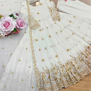 May include: A white Anarkali dress featuring gold embroidery and sequin embellishments. The garment includes long sleeves, a round neckline with a tie detail, and a flared skirt. A matching white dupatta with gold trim and floral accents is included.
