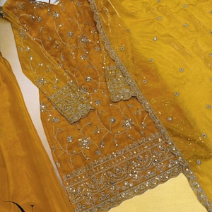 May include: Mustard yellow embroidered formal wear set. The set includes a long-sleeved tunic with intricate silver embroidery and sequin embellishments, a matching dupatta, and a skirt. The outfit is displayed on a wooden hanger.