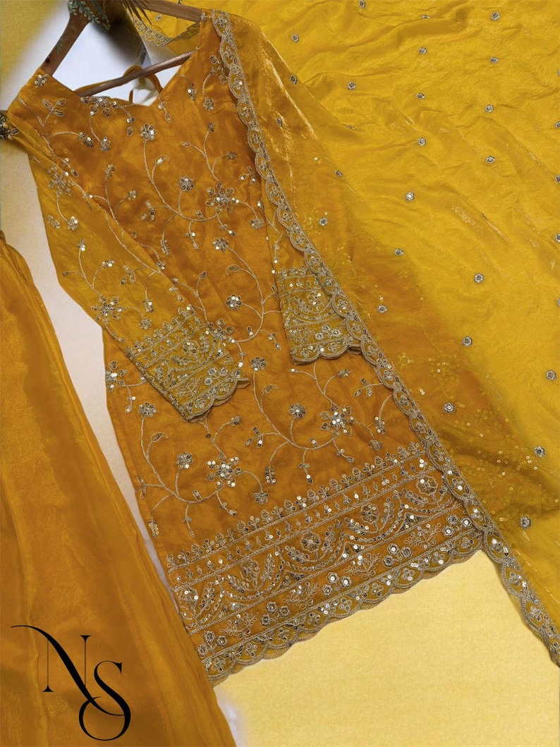 May include: Mustard yellow embroidered formal wear set. The set includes a long-sleeved tunic with intricate silver embroidery and sequin embellishments, a matching dupatta, and a skirt. The outfit is displayed on a wooden hanger.