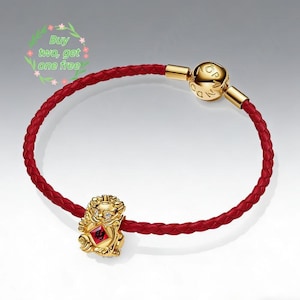 May include: A red braided leather bracelet with a gold charm. The charm is a detailed figure with a red and gold design. The bracelet has a gold clasp. The image also contains the text "Buy two, get one free".