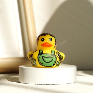 May include: A yellow rubber duck with black eyes and an orange beak. The duck is decorated with a green overall design and floral patterns. It is displayed on a white circular pedestal.